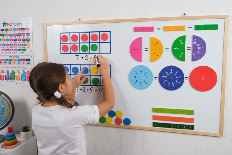 Little girl standing in math classroom near white magnetic board, smiling, solving math problems using frames, counters and fractions.