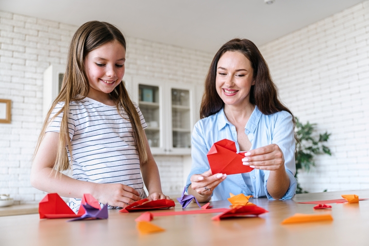 Smiling mother and daughter creating origami hearts out of red paper, sitting at table in dining room.