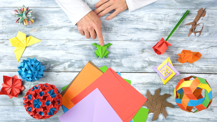 Origami folding lesson. Math student pointing at origami frog with finger. Collection of beautiful origami figurines on wooden table. Traditional origami paper folding.
