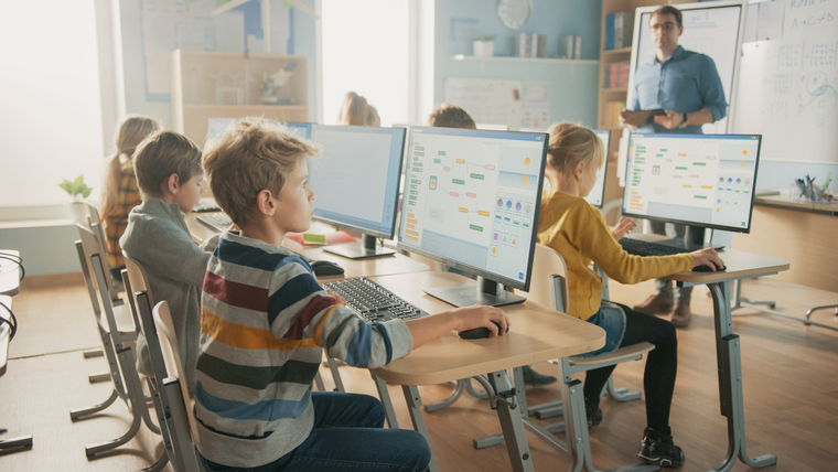 Elementary students learning computer programming in a modern classroom with desktop monitors, guided by a teacher at the front.