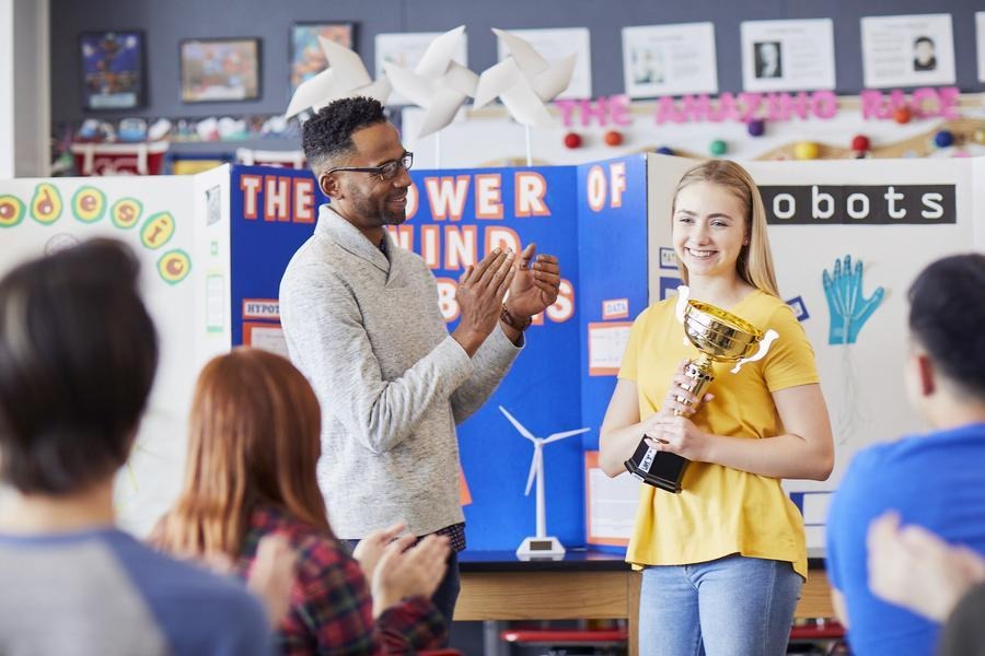 Student holding a trophy while teacher and classmates applaud at a school event.