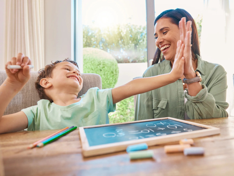 A woman and a child sit at a table, using chalk to write on a chalkboard in a bright, inviting room.