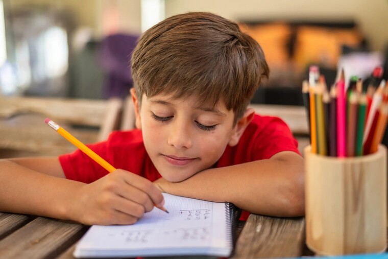 A young boy sits at a table, focused on writing in a notebook with a pencil.