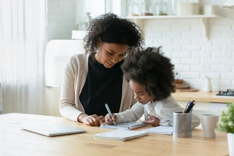 A mother and daughter collaborate on a school assignment at a table, surrounded by books and stationery.