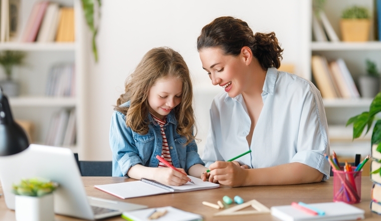 A woman and a child sit at a desk, focused on a laptop, engaged in a learning activity together.
