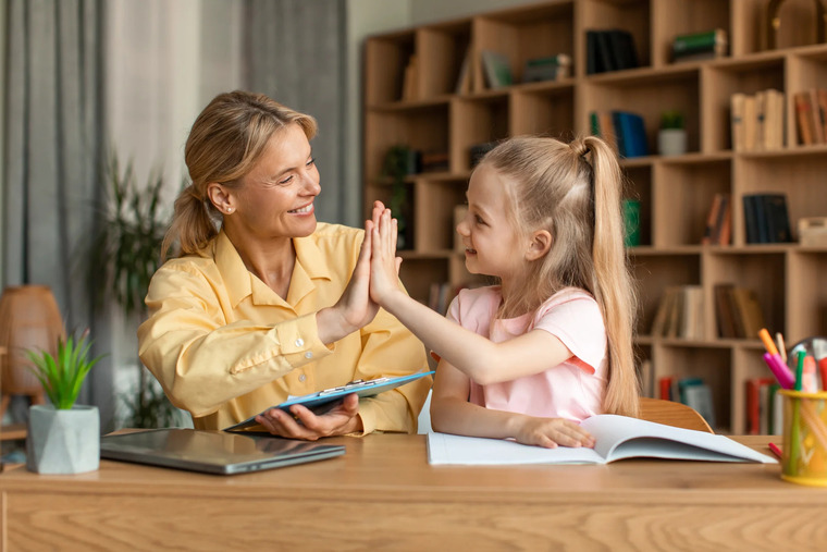A woman and a little girl are seated at a desk, both with their hands raised in excitement.