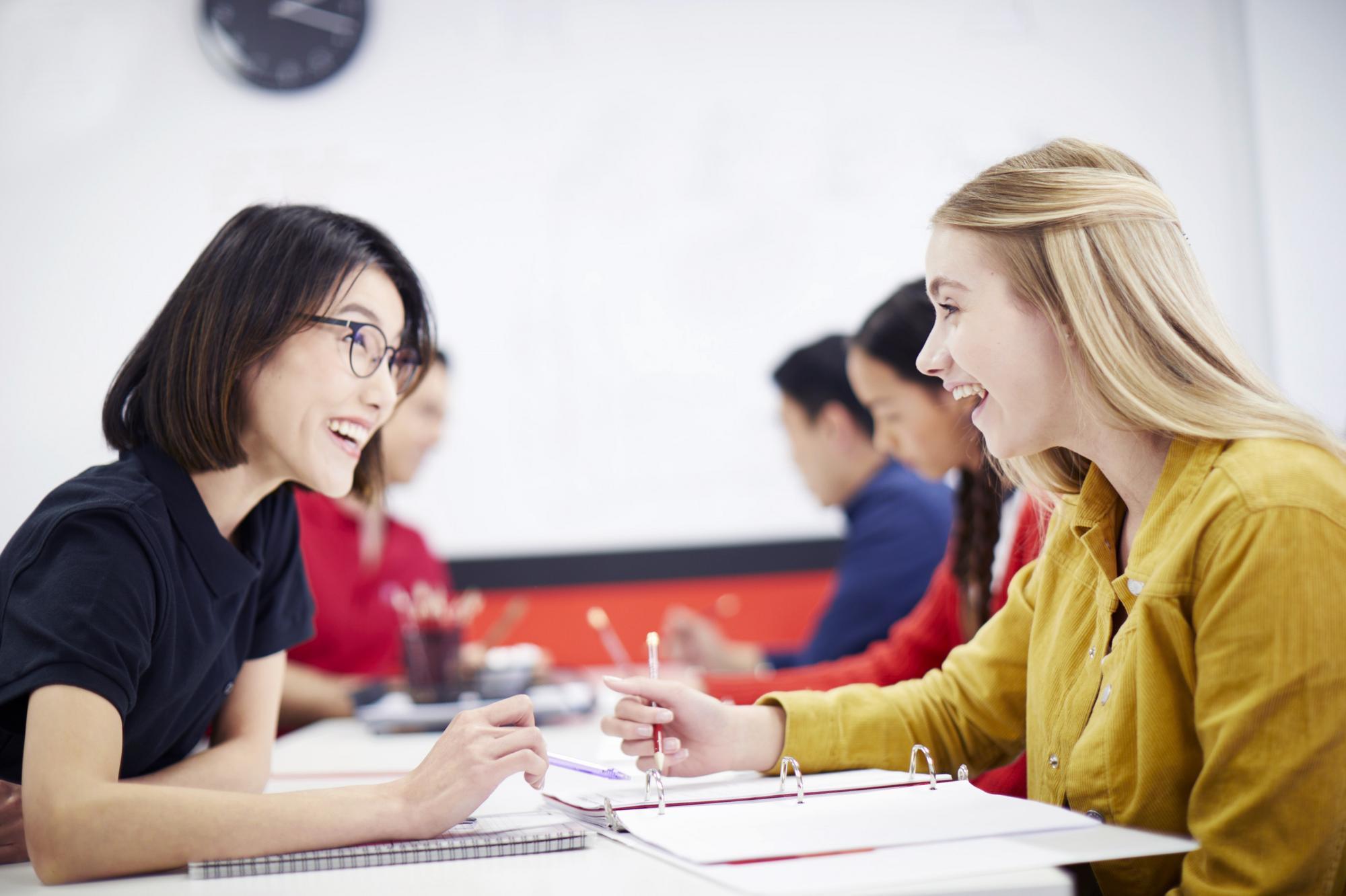 Two young women engaged in conversation in a classroom setting, surrounded by desks and educational materials.