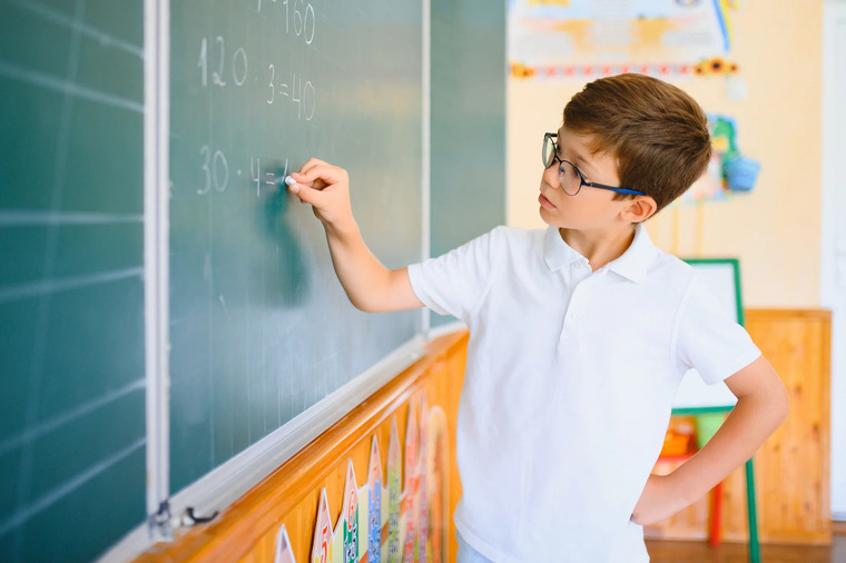 A boy wearing glasses is writing on a blackboard with chalk, focused on his task.