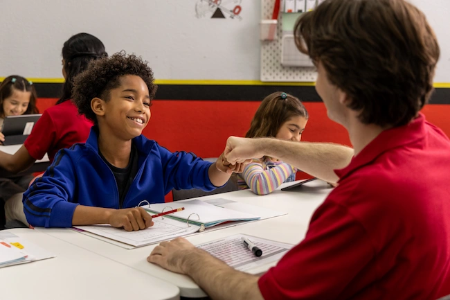 A man and a boy sit together at a classroom table, engaged in conversation and learning.