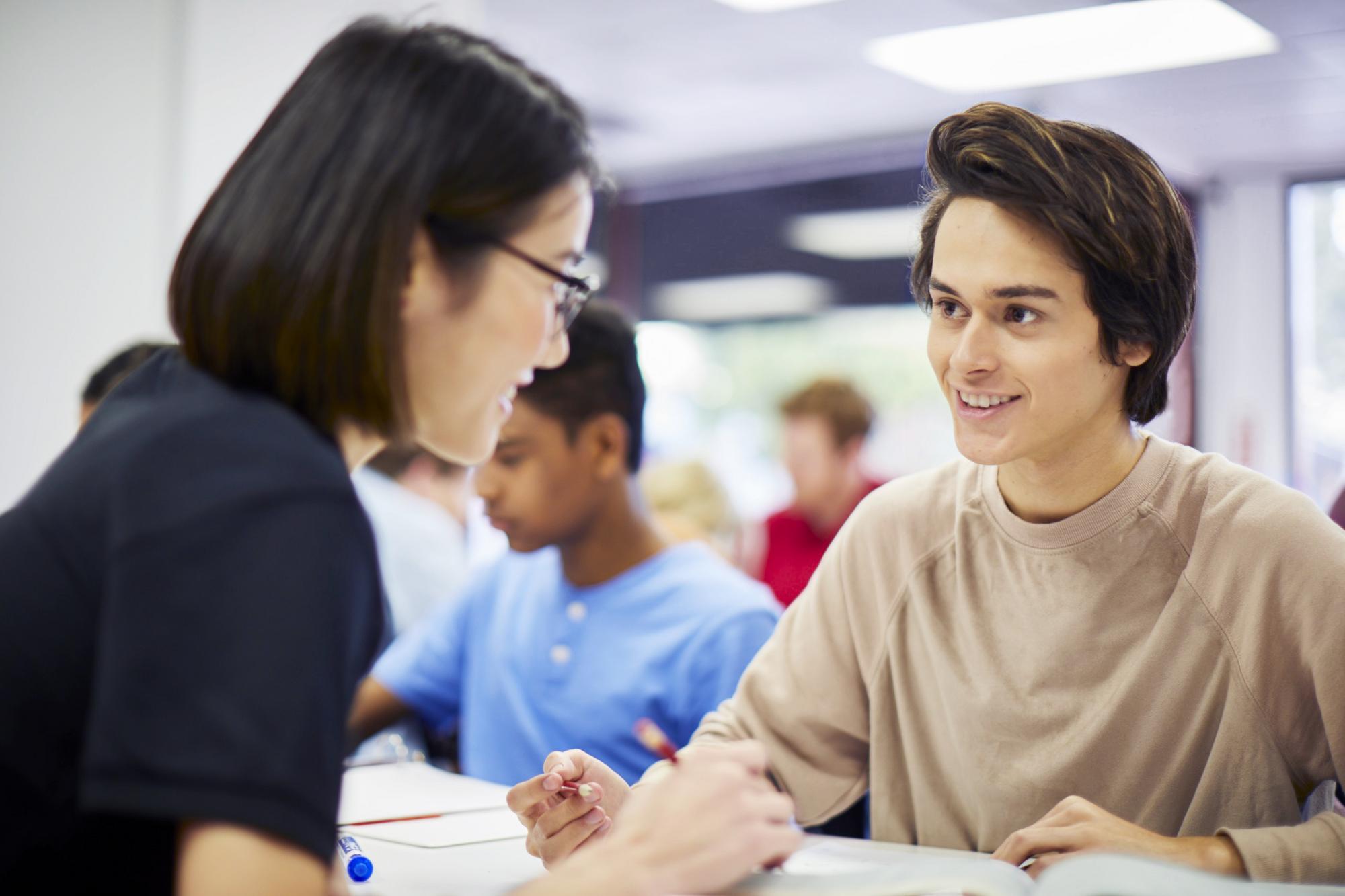 A young man and woman engaged in discussion at a classroom table, with books and notebooks in front of them.