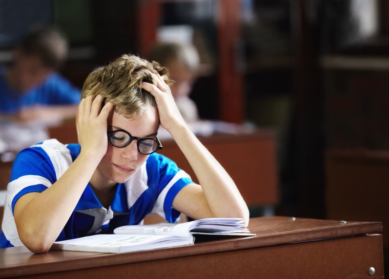 A boy with glasses sits at a desk, resting his head in his hands, appearing stressed or deep in thought.