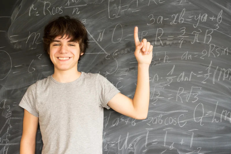 A young man gestures towards a chalkboard displaying various mathematical formulas and calculations.