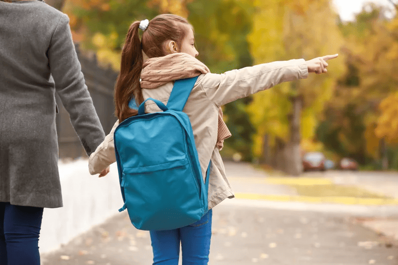A woman and a child walking hand in hand down a city street, surrounded by buildings and greenery.