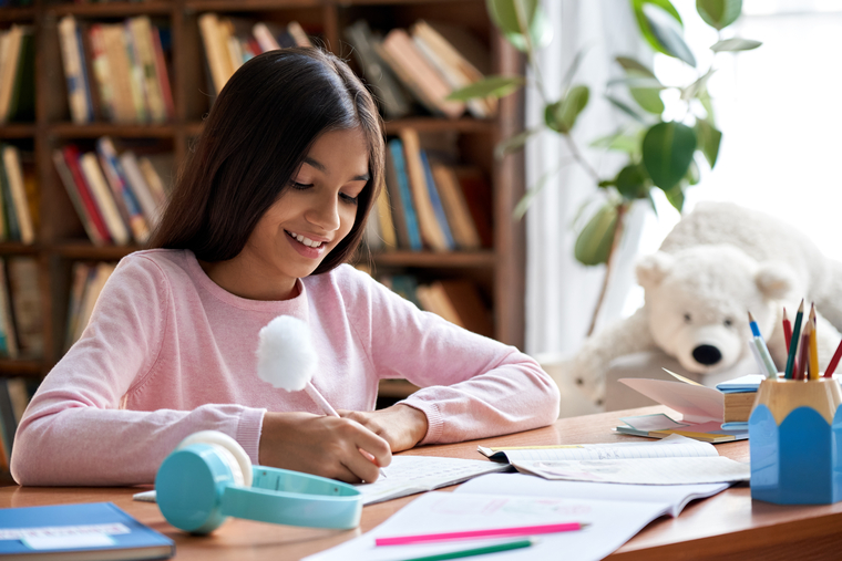 A girl sits at a desk, writing with a pen on paper, focused on her task.