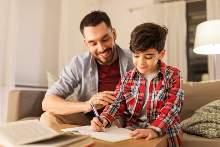 A man and a boy sit together on a couch, focused on writing in notebooks.