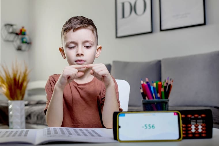A boy sits at a table, reading a book while holding a phone in his other hand.