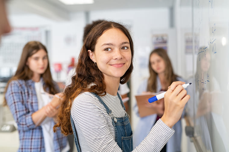 A girl writes on a whiteboard while other students observe and engage in a classroom setting.