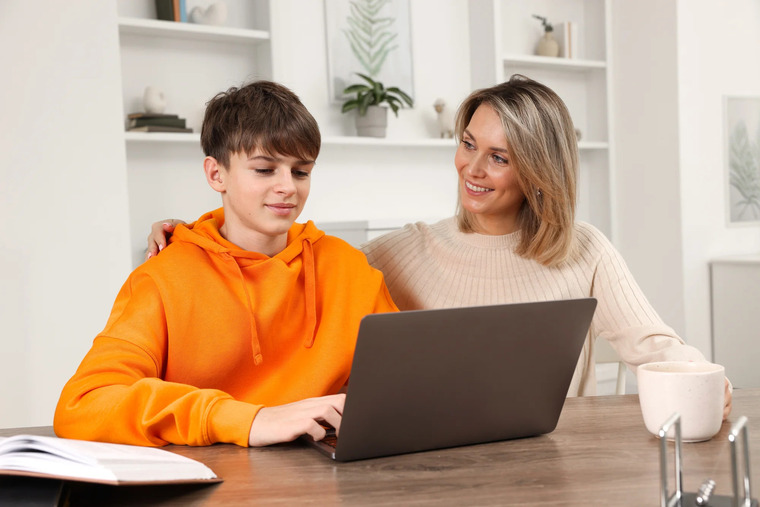 A woman and a boy are seated at a table, both looking at a laptop, sharing a learning experience.