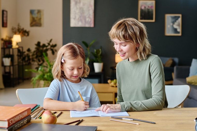 A woman and a child sit at a table, engaged in writing with a pen and paper.