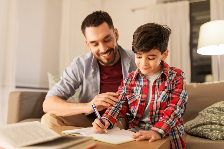 A man and a young boy are seated on a couch, engaged in writing activities with notebooks.