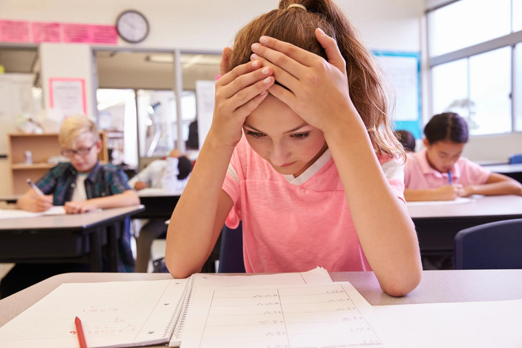 A girl is seated at a desk, her head resting in her hands, suggesting feelings of anxiety or exhaustion.