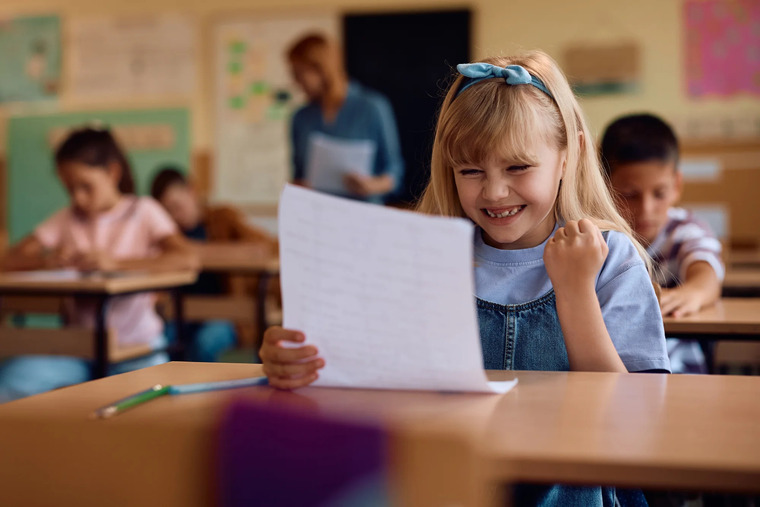 A cheerful little girl reads a paper in a classroom, showcasing her engagement and enthusiasm for learning.