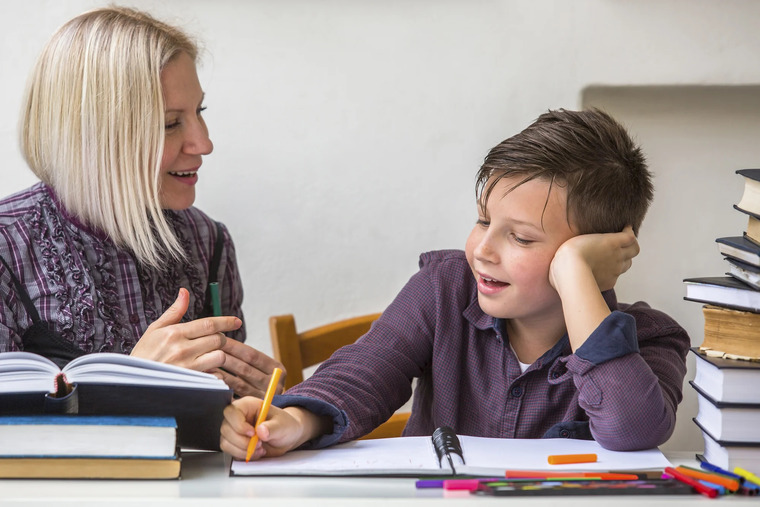 A woman and a boy sit at a table, surrounded by open books, engaged in reading and discussion.