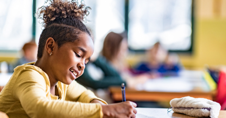 A young girl sits at her desk in a classroom, focused on writing in her notebook.