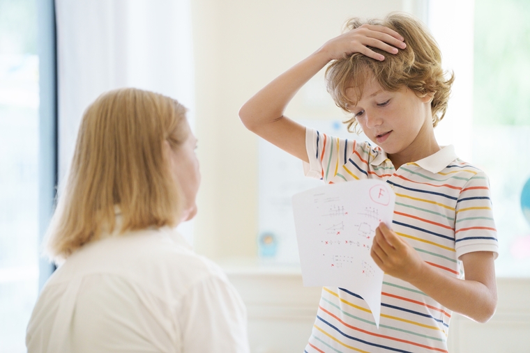 Worried child holding a test marked with a failing grade, standing in front of his mom
