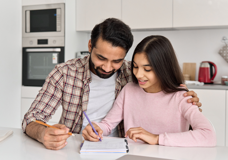 Father helping his daughter with homework at the kitchen table, both smiling as they work together in a supportive and positive atmosphere