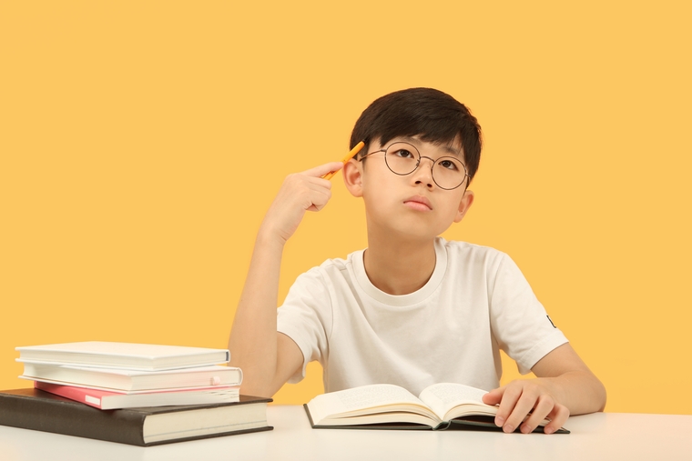 Boy wearing glasses sitting at a desk with books, holding a pencil to his temple and thinking while looking up, symbolizing reflection and problem-solving in math learning