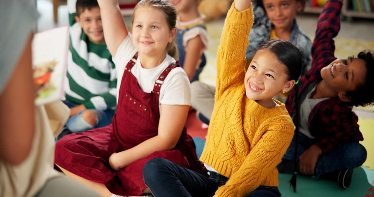 Several children seated on the floor, hands up, showing enthusiasm and readiness to answer questions in a classroom setting.
