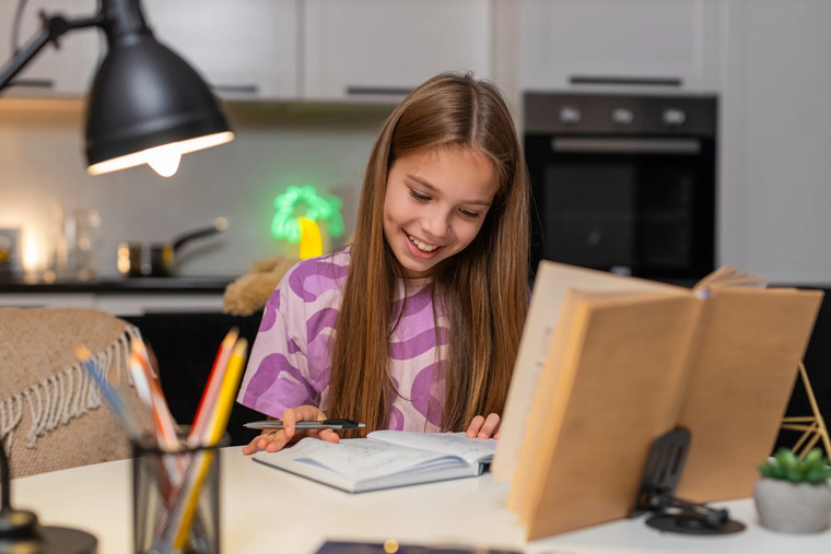 A cheerful girl at a table, smiling while surrounded by a book and a laptop, indicating a moment of study or leisure.