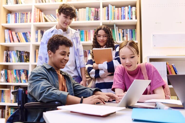 A group of students seated at tables in a library, focused on their laptops while studying and collaborating.