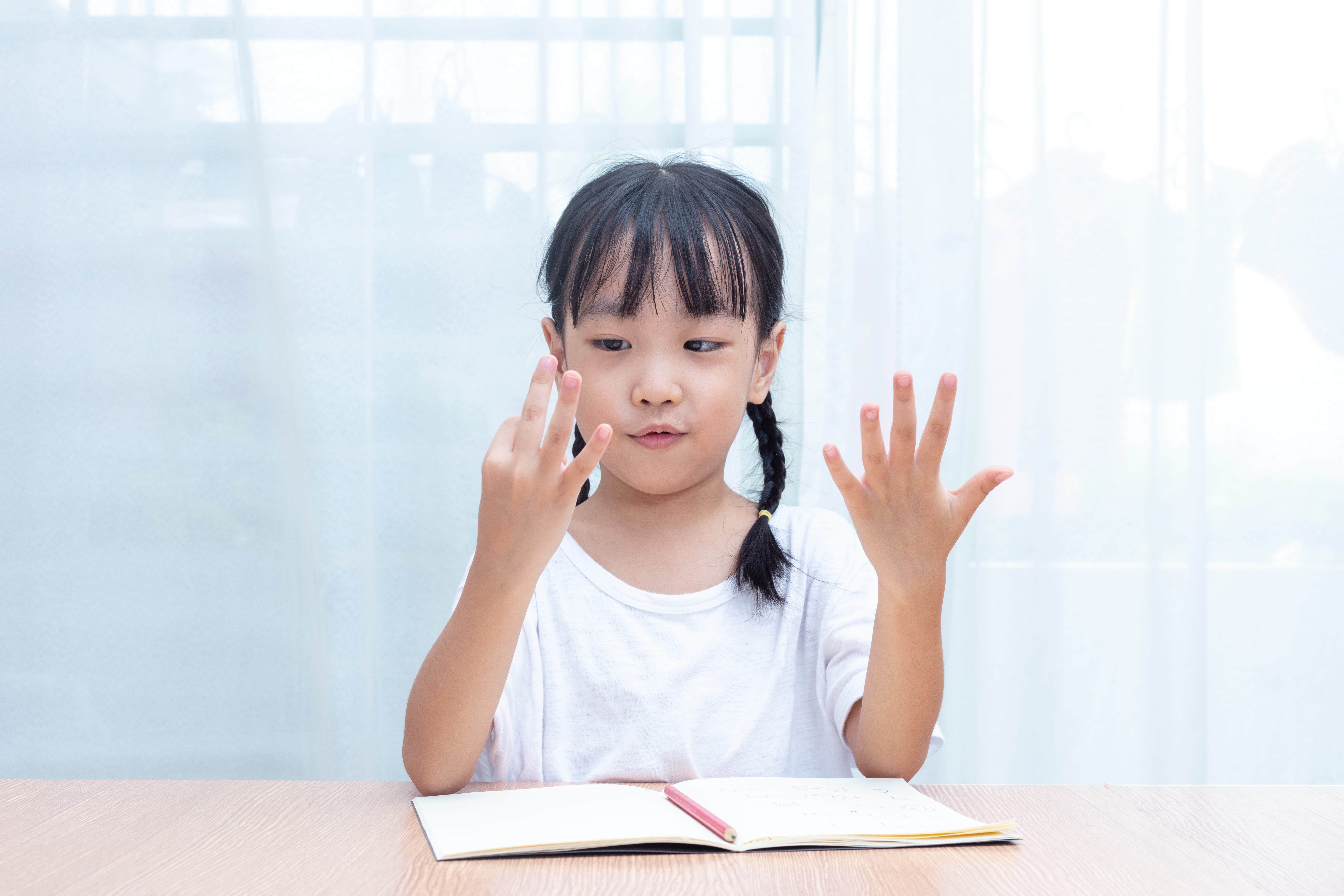 A girl counting with her fingers while she sits at the table with an open notebook.