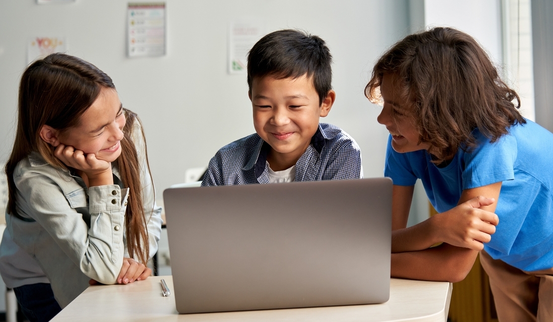 Three elementary school students in a math class smiling and working together on a laptop.