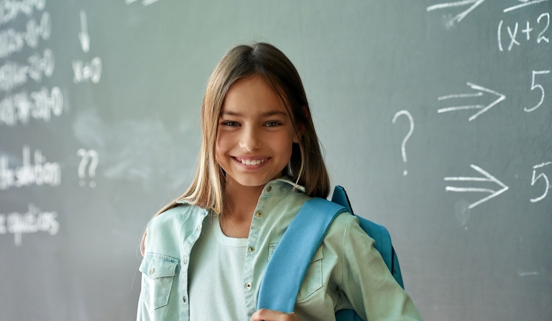 Happy cheerful smiling cute schoolgirl with backpack standing in math classroom on a chalkboard blackboard background.