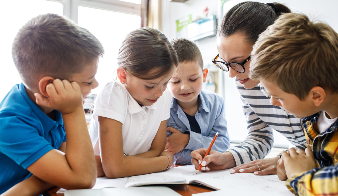 Tutor helping a group of young students with math work at a table, fostering engagement and collaborative learning in a classroom setting
