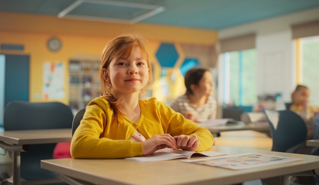 Smiling elementary school girl sitting at a classroom desk with workbook