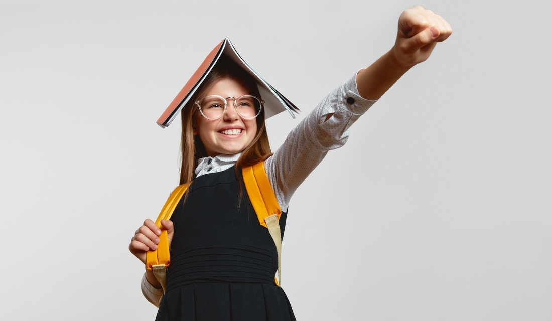 Confident young girl in school uniform and backpack smiling with a book on her head like a superhero cape, raising her fist in a heroic pose