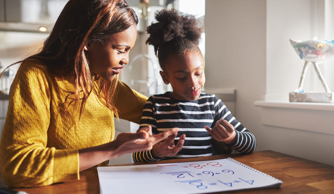 Mother helping her daughter with math homework at the kitchen table, both using fingers to solve problems in a workbook