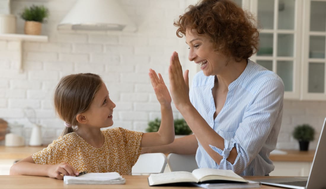 A woman and a young girl sit at a table, both raising their hands in a gesture of excitement.