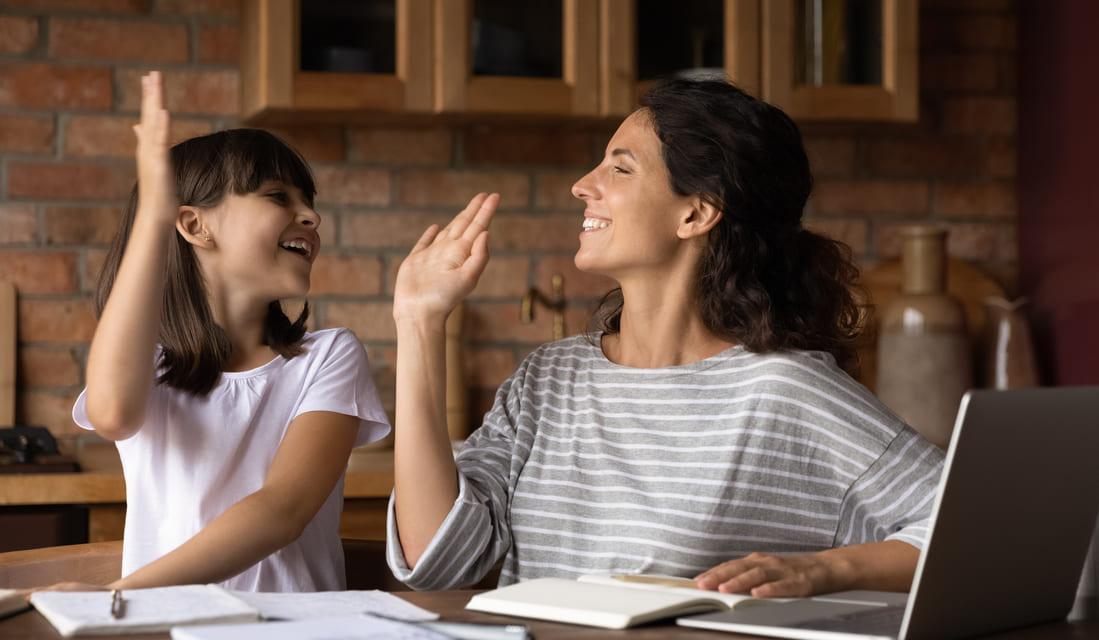 A woman and her daughter sit at a table, both raising their hands in a cheerful gesture.