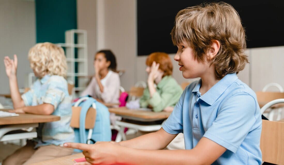 A boy sitting at a classroom desk with his hand raised, indicating he wants to answer a question.