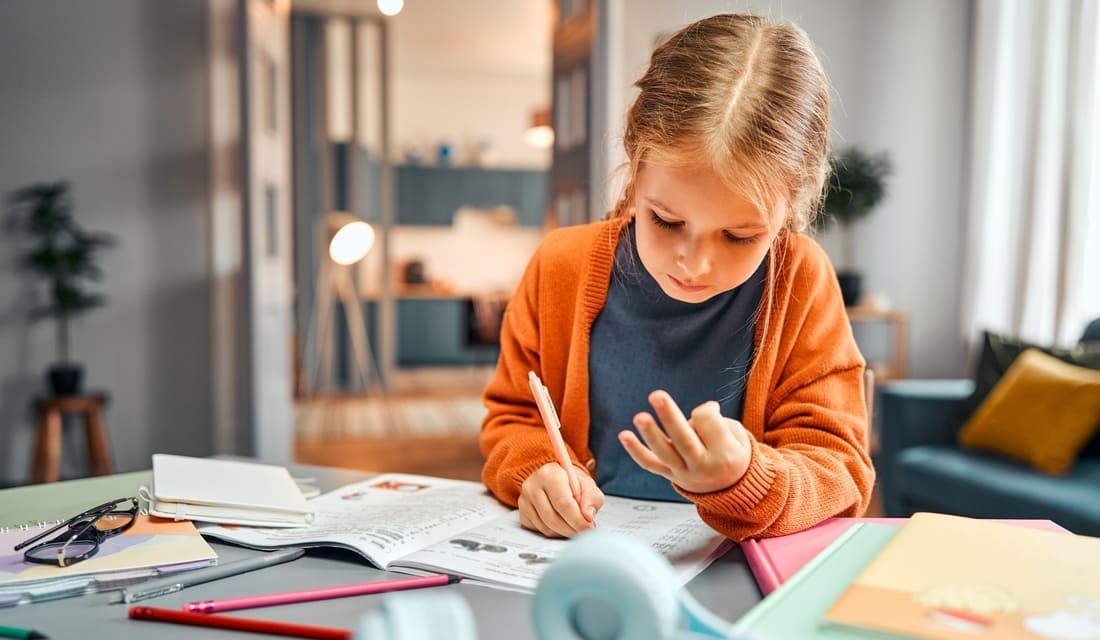 A young girl sits at a table, focused on her notebook while holding a pencil, engaged in writing or drawing.
