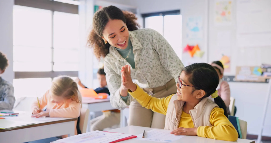 A teacher assists a young student with their work in a bright, engaging classroom setting.