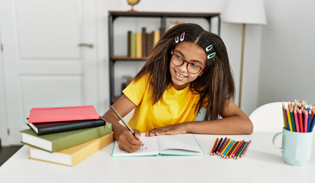 A young girl sits at a table, focused on her notebook, surrounded by colorful pencils.