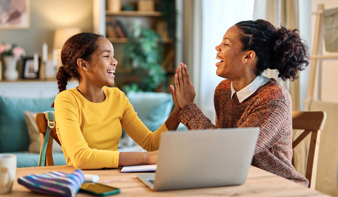 A mother and daughter celebrate together at a table, high-fiving while engaged with a laptop in front of them.