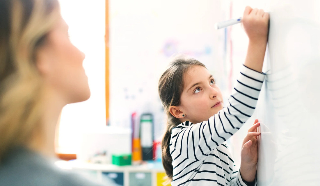 A mother assists her young daughter as they write on a whiteboard, fostering a supportive educational environment.