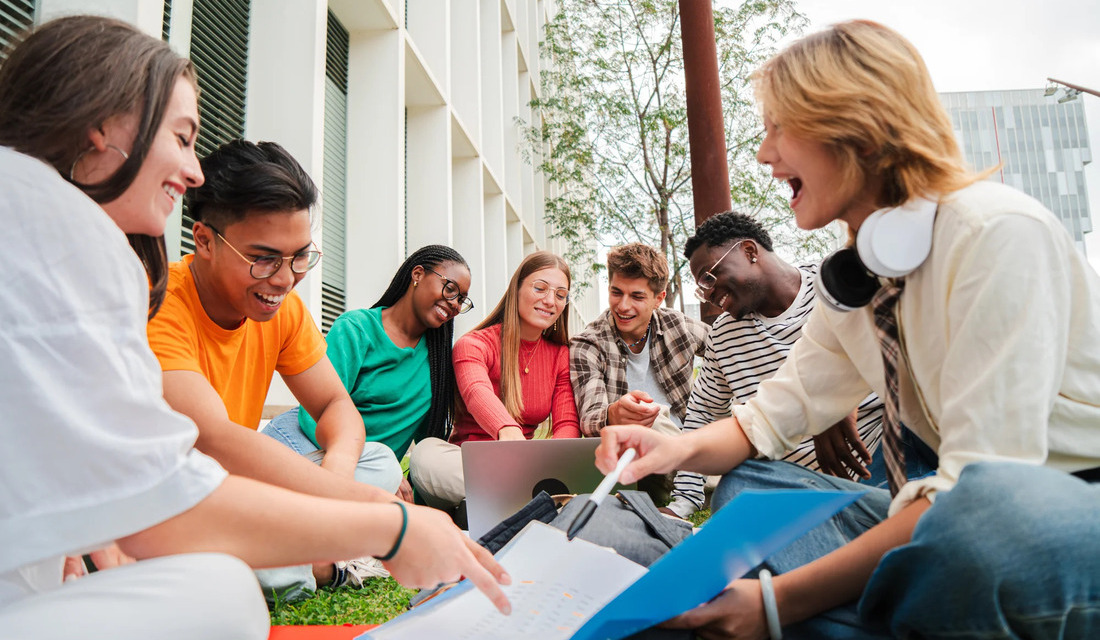 A group of students sitting on grass, collaborating on a laptop in a sunny outdoor setting.